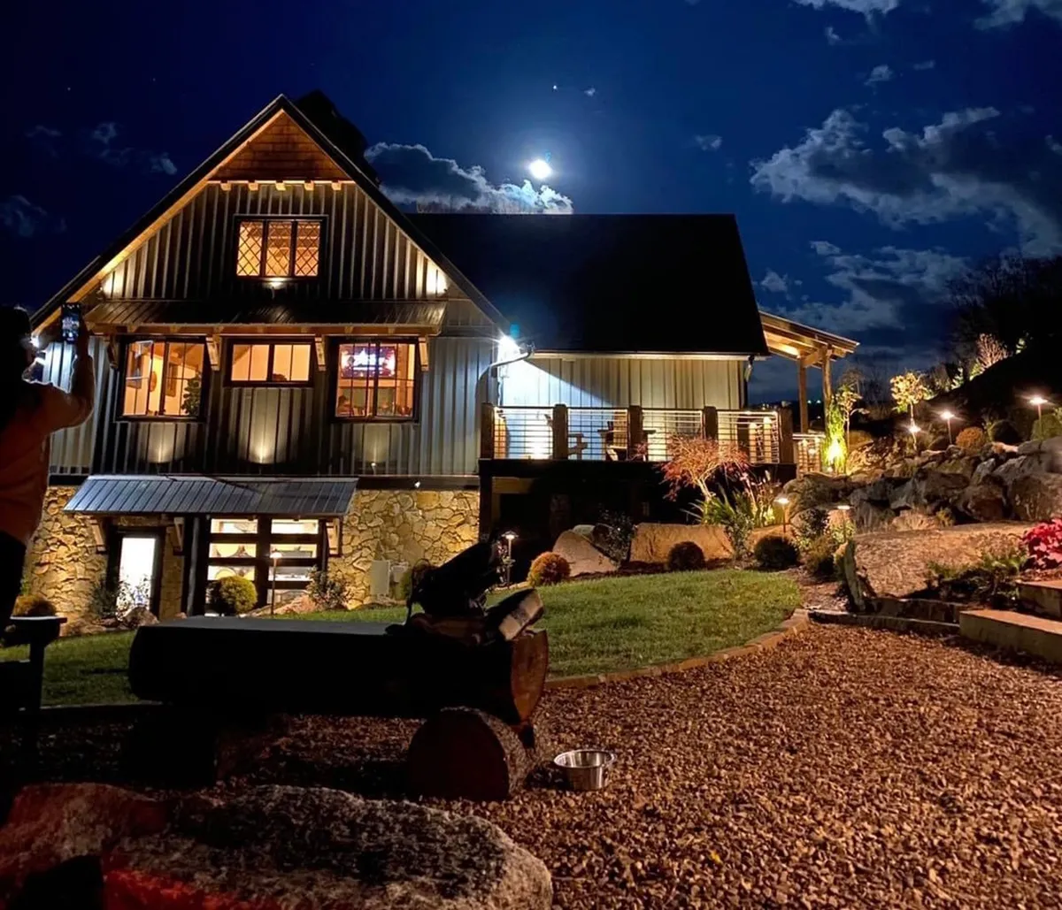 An atmospheric exterior shot featuring the illuminated house at night, a glowing moon overhead, lush garden, and a cozy fire pit area. The scene suggests evenings spent outdoors, enjoying tranquil moments and group activities under the stars, with pets close by.