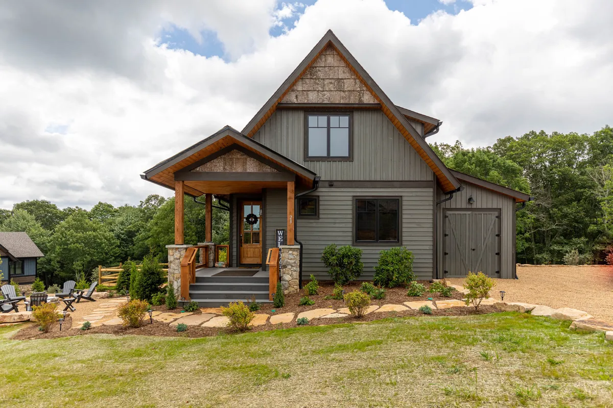 The image features the exterior of the property, showcasing a welcoming front porch with wooden accents surrounded by a landscaped garden. A stone pathway leads to the porch, and the setting is bathed in warm, inviting light, suggesting an ideal spot for relaxing and enjoying seasonal outdoor gatherings. The vantage point allows for the appreciation of sunsets from the porch, emphasizing the connection to the surrounding natural beauty.