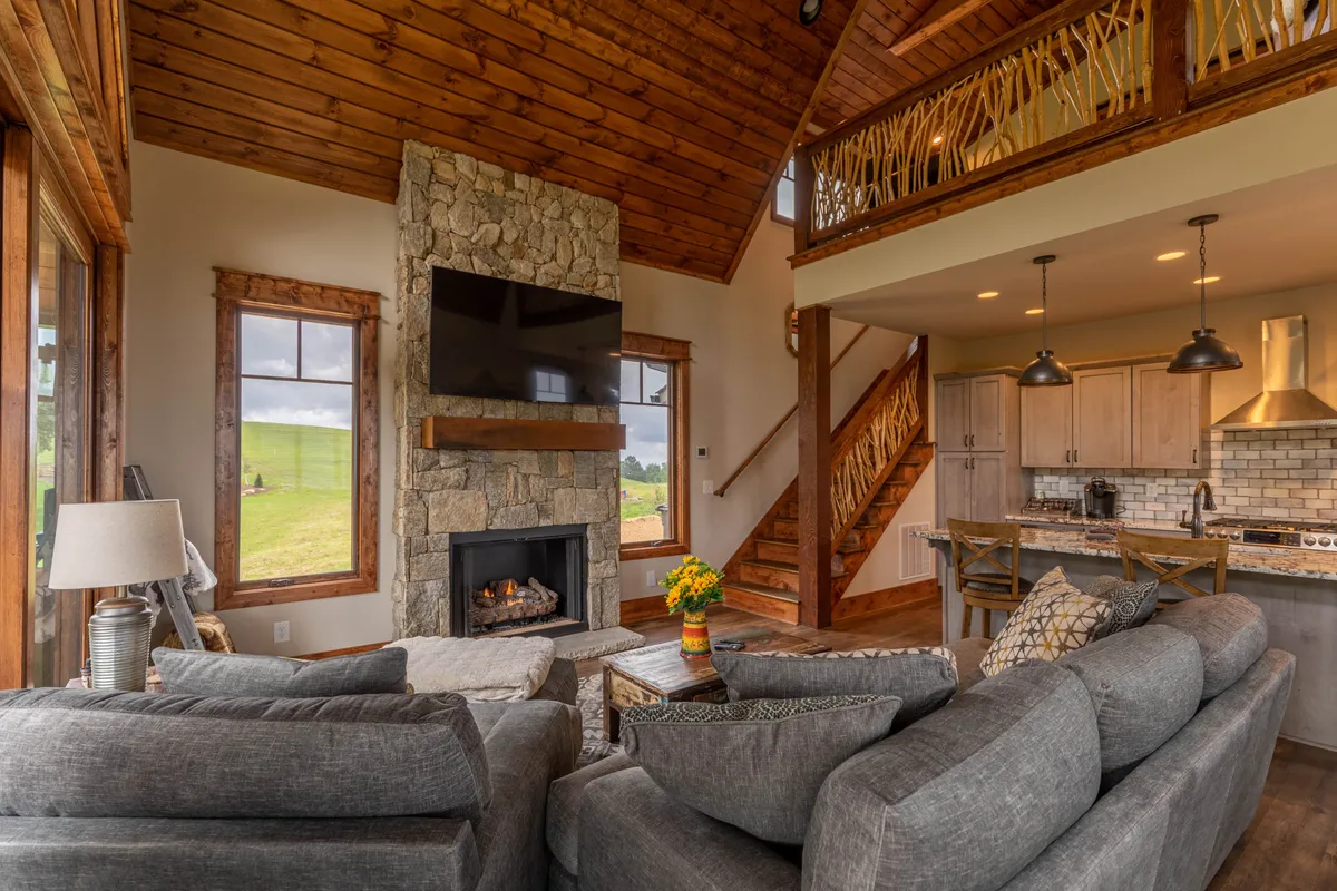 A warm, open-concept living area featuring a prominent stone fireplace as the focal point, a large sectional couch perfect for gatherings, a wooden staircase adding architectural interest, and a kitchen island visible in the background. The space radiates coziness and encourages togetherness.