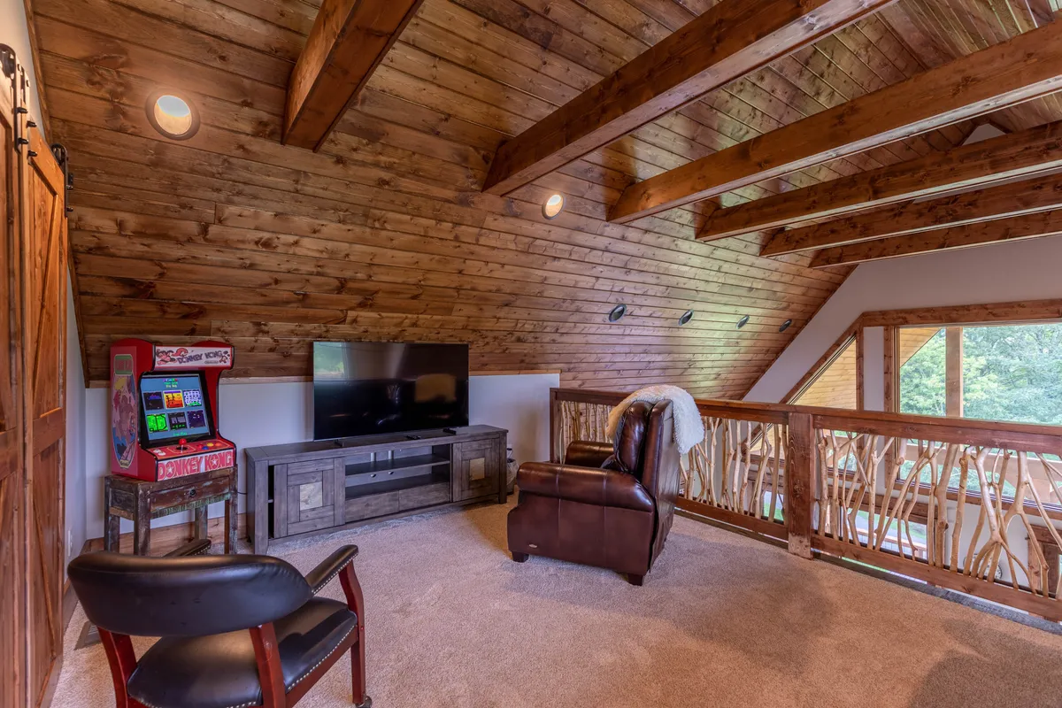 A welcoming upstairs loft area showcasing wooden ceiling beams, a television, a leather recliner, and, most notably, a classic arcade machine beside the balcony railing. The setting is casual, playful, and perfect for leisure activities or family fun.