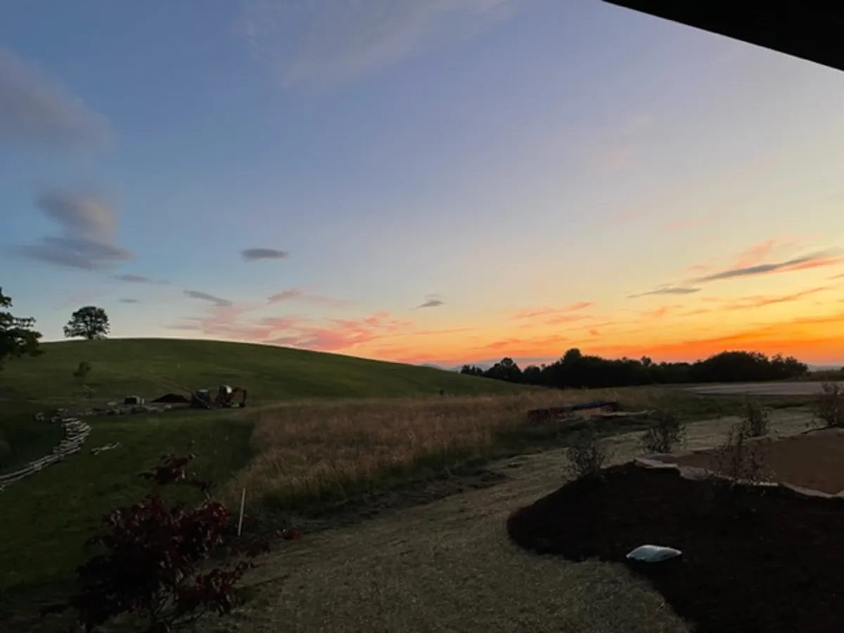 A panoramic view of rolling hills and distant trees under vibrant sunset hues. The tranquil scene captures the serenity of the surrounding landscape, suggesting opportunities for stargazing, nature walks, or simply soaking in the mountain air and quiet moments.