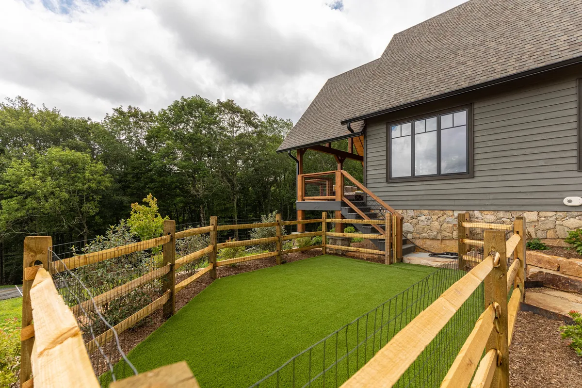 A bright, welcoming backyard scene featuring a green artificial grass area bordered by a wooden fence, with steps leading up to the entrance. The yard's spaciousness and secure fencing make it ideal for pets and family activities.