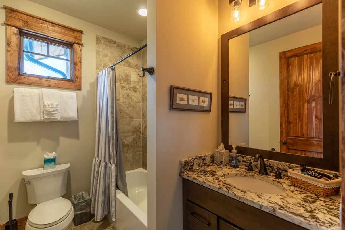 A modern bathroom with a granite countertop, a large mirror, and a tiled shower area, complemented by elegant wooden accents and framed artwork. The space feels bright, fresh, and soothing, ideal for relaxing after a day outdoors.
