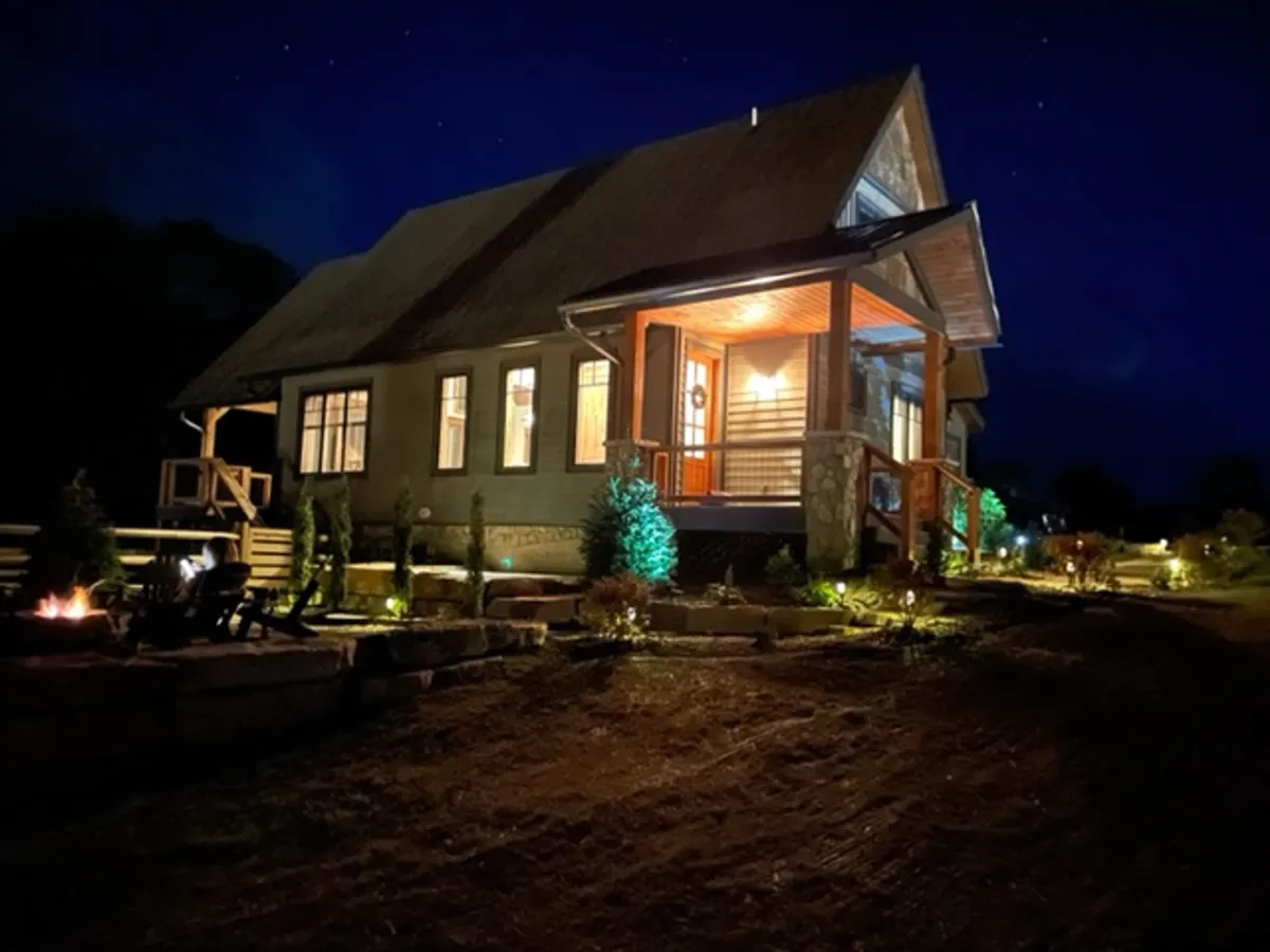 A welcoming exterior shot featuring a front porch, landscaped garden, and an inviting fire pit surrounded by decorative lighting. The cozy setting is ideal for gathering in the evenings, stargazing, and roasting marshmallows by the fire.