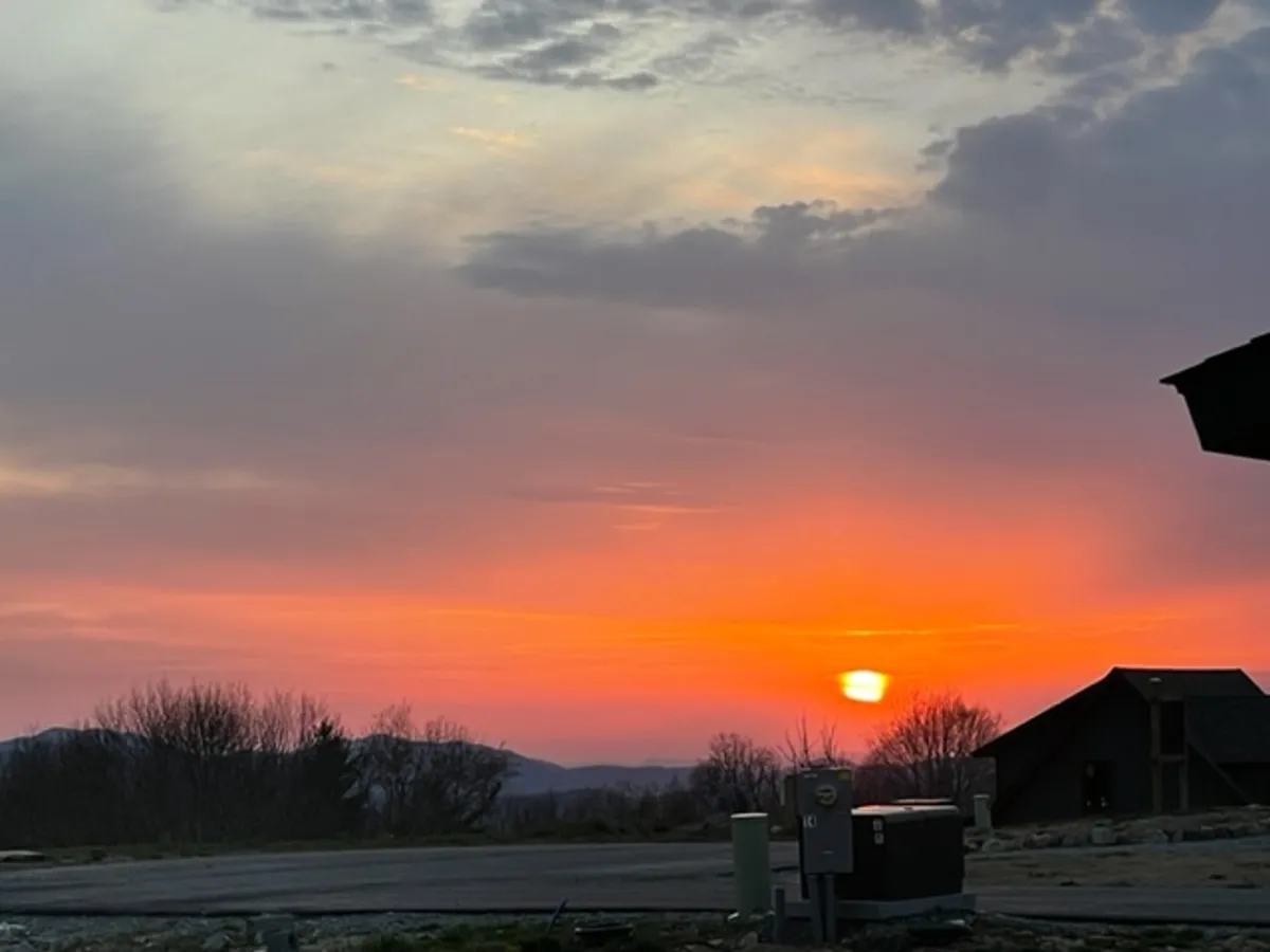 A captivating view of a colorful sunset over distant mountains, with silhouettes of trees and the outline of a building in the foreground. The ambiance is peaceful and expansive, inviting reflection and relaxation.