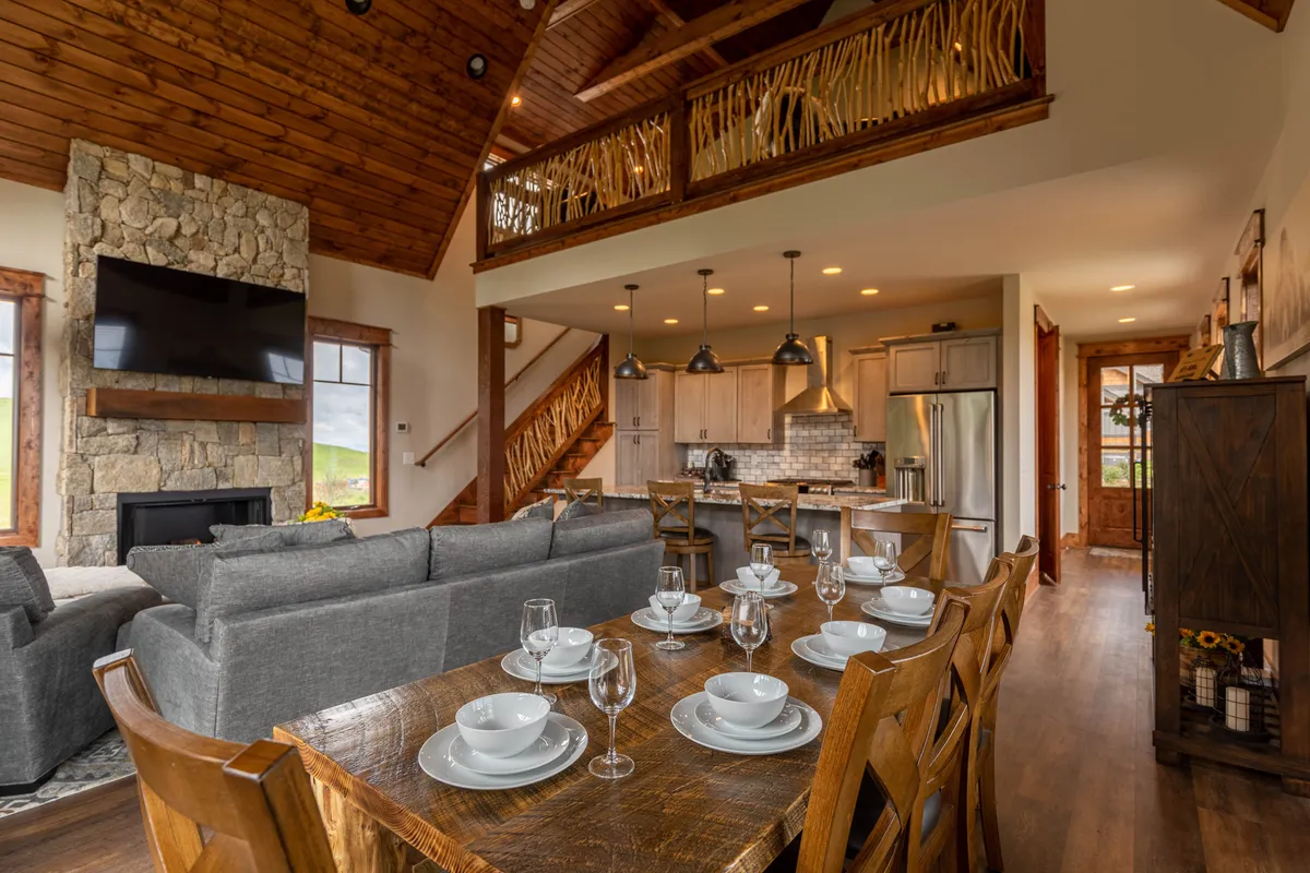 An inviting open-concept space that combines a stone fireplace, a large dining table, and an open kitchen with wooden beams overhead. The layout naturally supports family meals, entertaining guests, and celebrating together, all in a warm, communal setting.