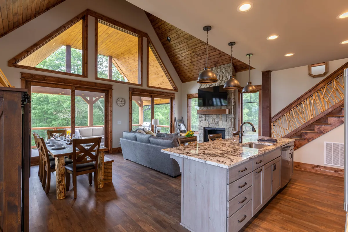 An inviting interior view featuring a granite-topped kitchen island, wooden dining table, large windows flooding the room with daylight, and a stone fireplace, all arranged to maximize social interaction and comfort.