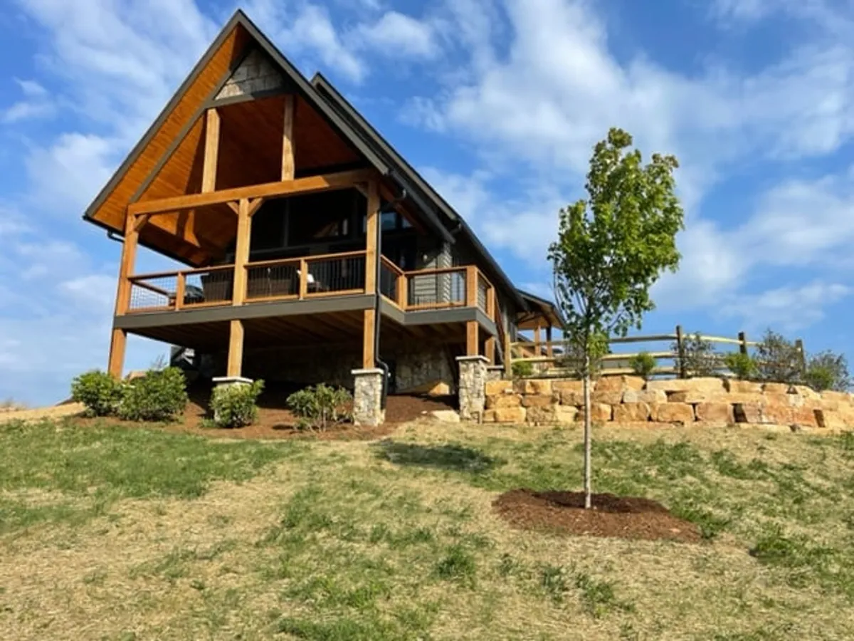 The image showcases a stunning wooden mountain lodge with a spacious porch, set against lush greenery and anchored by a rustic stone wall. The porch offers sweeping views perfect for sunsets, while the setting radiates warmth and tranquility—ideal for gathering with family and friends or unwinding in nature.
