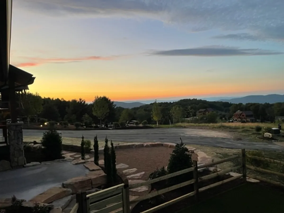 A wide-angle view featuring a sunset sky over a scenic mountain range, landscaped areas, and a meandering pathway. The mood is serene and majestic, inviting guests to imagine exploring the lodge’s beautiful natural surroundings.