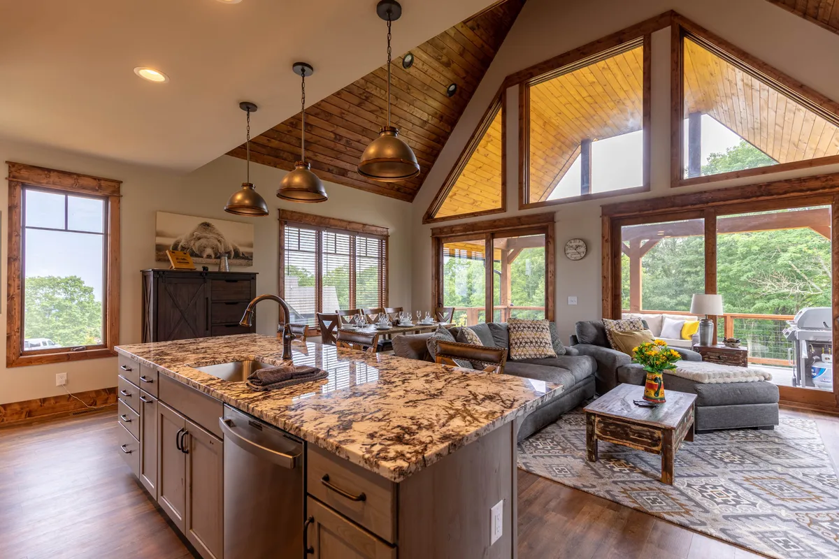 A bright, airy view of the kitchen and living space with a spacious kitchen island, large windows streaming in natural mountain light, a dining table, and a comfortable seating area. The layout emphasizes seamless flow between cooking, dining, and relaxing.