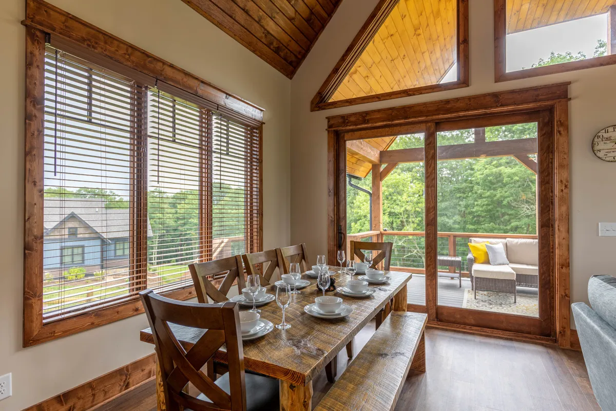 The interior dining area boasts large windows and a sliding glass door that opens to an outdoor seating space. The room is filled with natural light, providing clear views of the surrounding nature and creating a seamless connection between the interior and exterior environments. The setting is warm and inviting, ideal for sharing meals while enjoying beautiful vistas.
