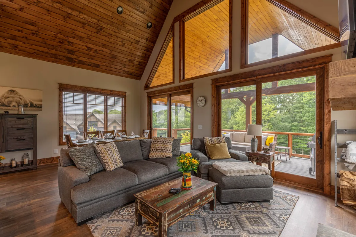 A spacious, cozy living area featuring large windows that invite natural light, a comfortable sofa with decorative pillows, a wooden coffee table adorned with vibrant flowers, and views that hint at the outdoor landscape. The seating arrangement is perfect for group gatherings, game nights, or relaxed conversations with family and friends.
