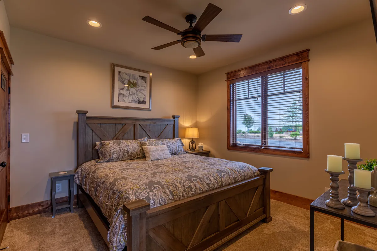 A tranquil bedroom featuring a large bed with a patterned bedspread, wooden furniture, and a window with blinds that allows natural light to stream in. Decorative artwork adds a touch of personality, while the overall scene radiates calm and comfort.